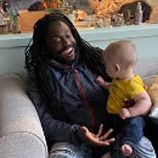 Mike Brown is sitting on a sofa interacting with a baby, who is on his lap.