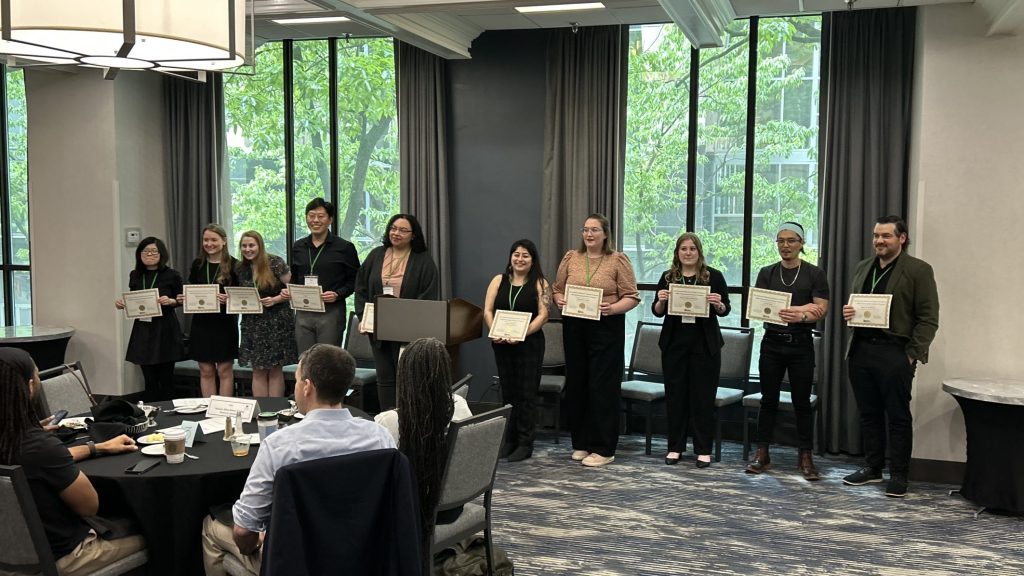 A group of NEFA students stand in a row at the front of the room, holding certificates