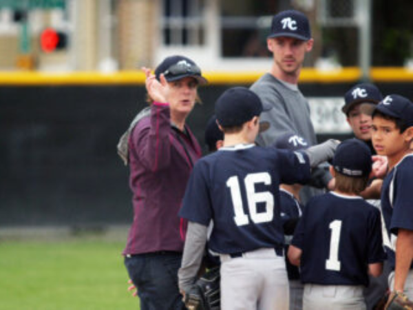 A coach, surrounded by young baseball players, gestures to the field.
