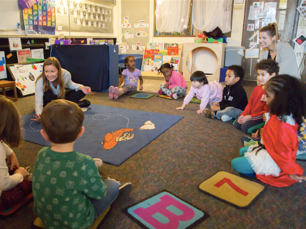 Students sit in a semi-circle facing a smiling teacher. Another teacher is seated in the circle.