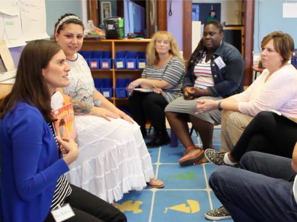 One teacher holds a picture book and talks with the other teachers gathered around.