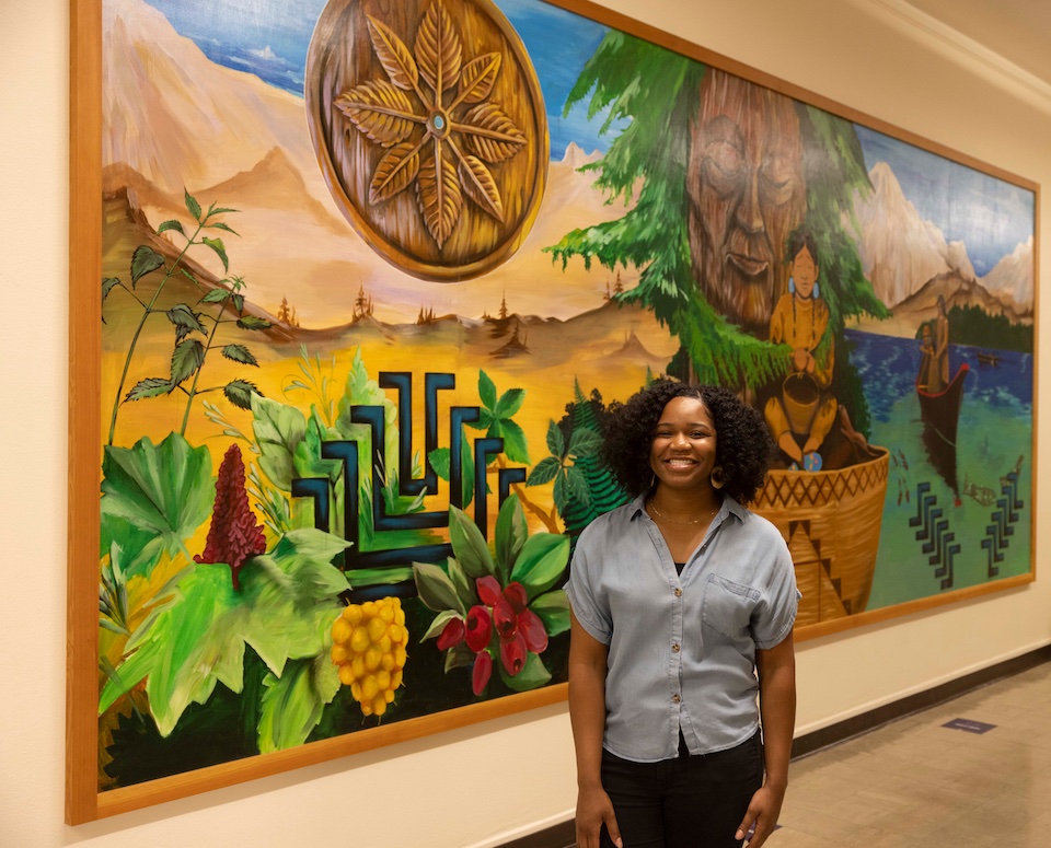 Doctoral candidate and Banks Center graduate assistant Jazmen Moore photographed near a mural by Indigenous artists Roger Fernandes and Toma Villa in Miller Hall.