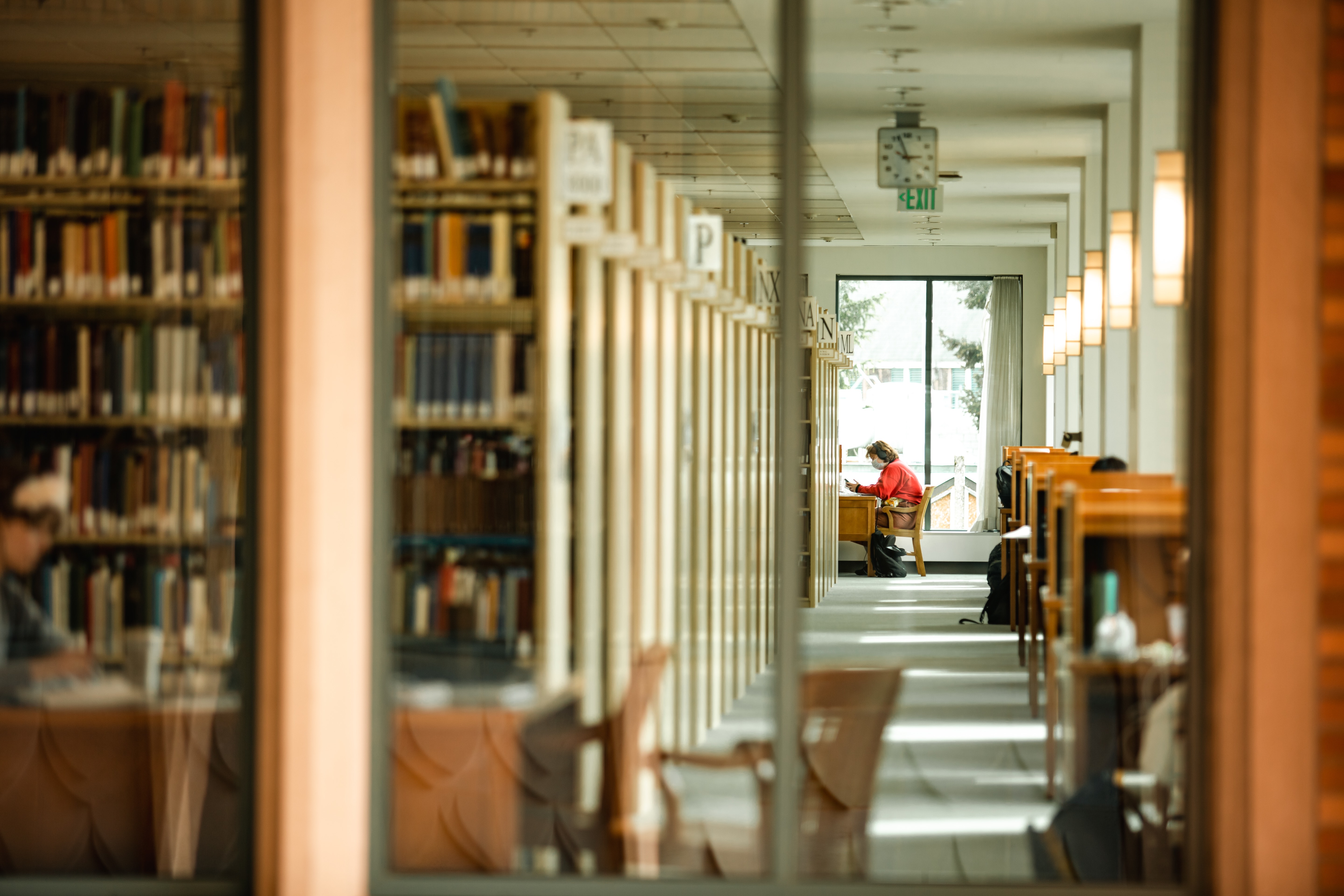 A student sitting in a library. Rows of book stacks are featured.